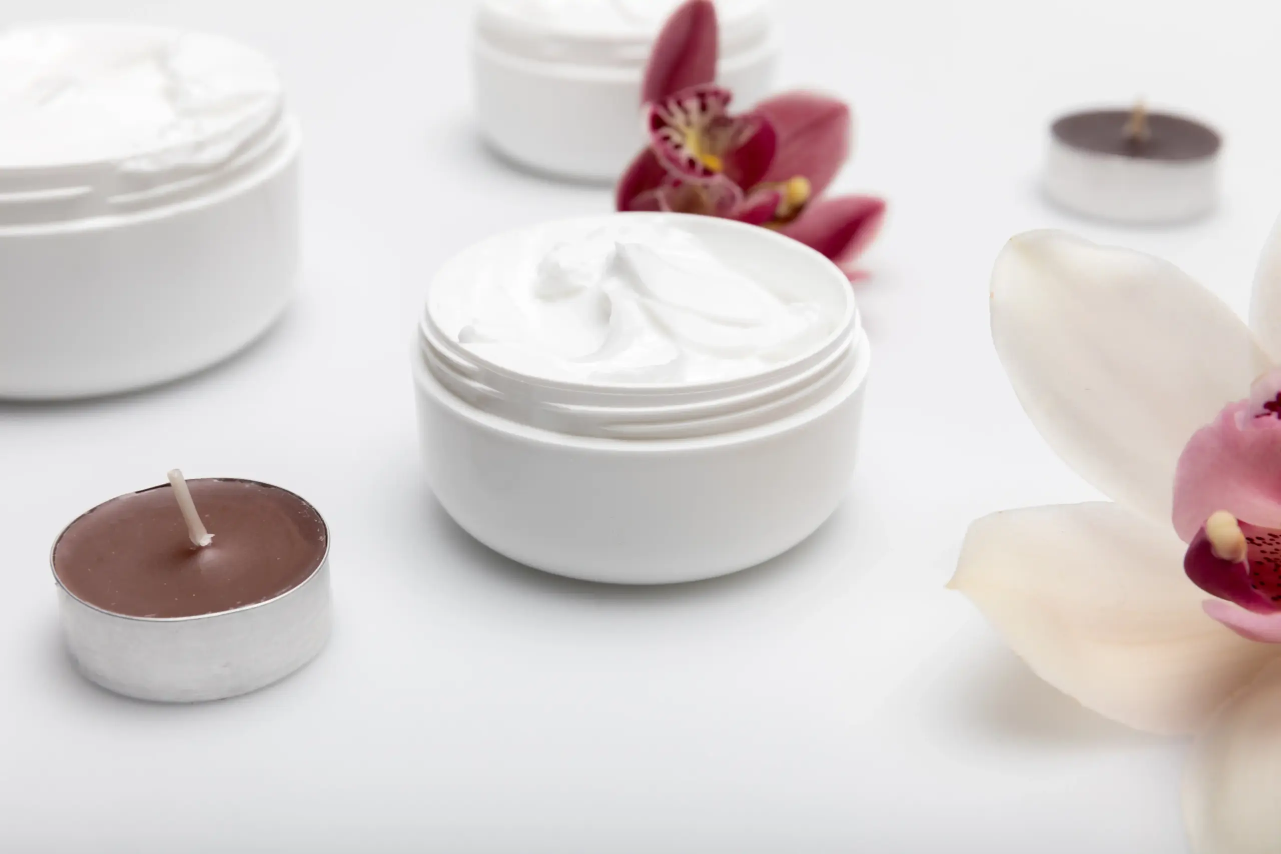 Krim pelembap alami dalam wadah putih dengan bunga anggrek dan lilin aromaterapi di latar putih, konsep perawatan kulit alami dan relaksasi spa.